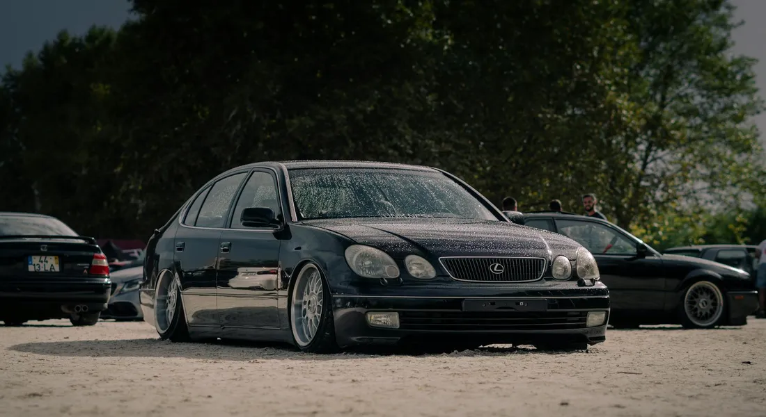 A lowered dark-colored sedan with white aftermarket wheels parked in an outdoor lot, with other cars and trees in the background.
