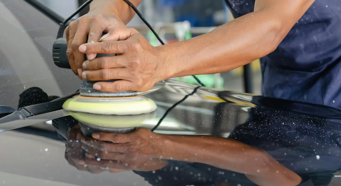 Detailer uses a foam-pad polisher on a car hood, reflecting surface as they work on removing paint overspray.