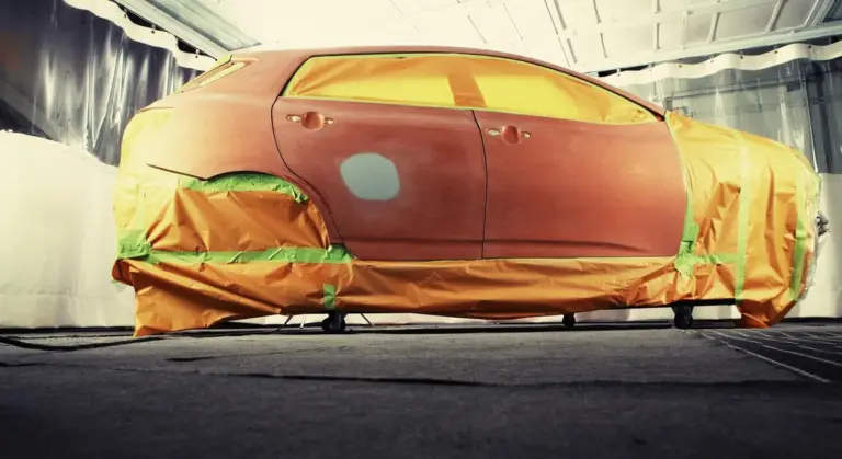 Car masked for paint prep inside a spray booth, with orange tape covering edges and patched panel areas.