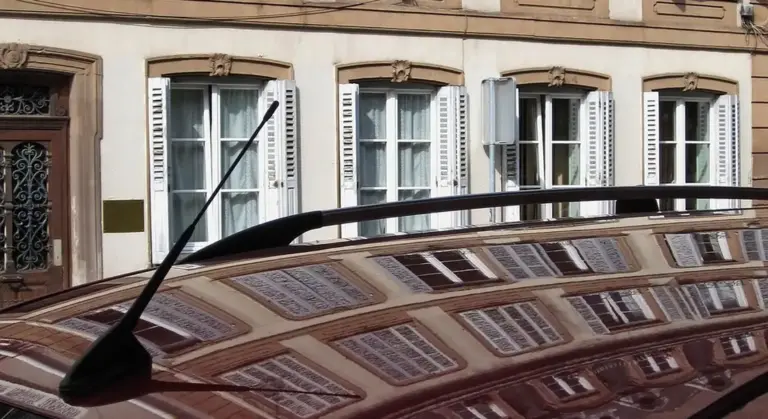 Close-up of a glossy car hood with a windshield wiper, reflecting the facade of a row of windows and buildings.