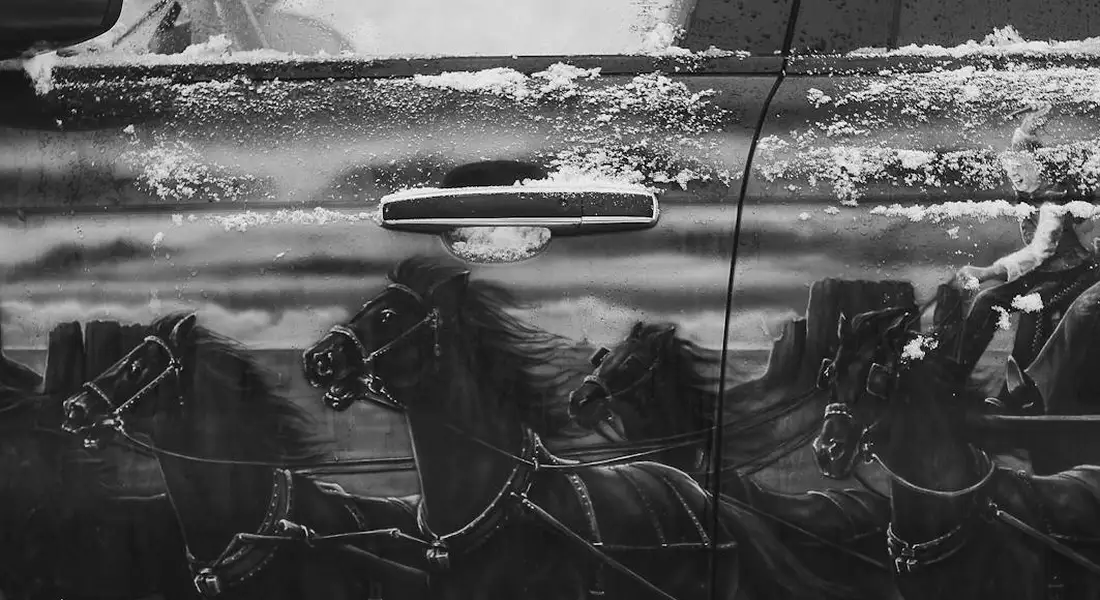 Black-and-white car door with a reflected image of horse-drawn carriages on its surface.