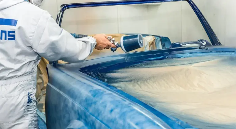 Technician in a white protective suit uses a spray gun to apply paint on a blue car inside a spray booth, with masked windows.