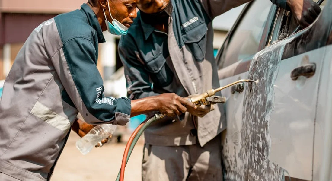 Two technicians in work uniforms remove spray paint splatter from a car's exterior panel, using a tool near the door and trim.
