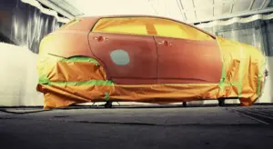 A car inside a spray booth masked with orange tape and protective paper, awaiting repaint to restore a like-new finish.
