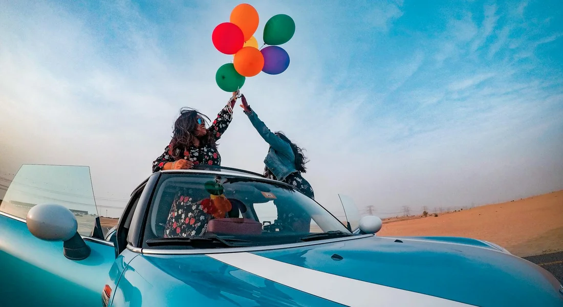 A blue car with two people on the roof holding colorful balloons in a desert, symbolizing the milestone of finishing the sanding stage before painting.