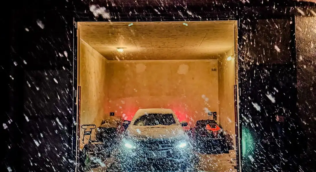 A car exits a dimly lit garage during a snowy night, headlights shining as snowflakes fall around it.