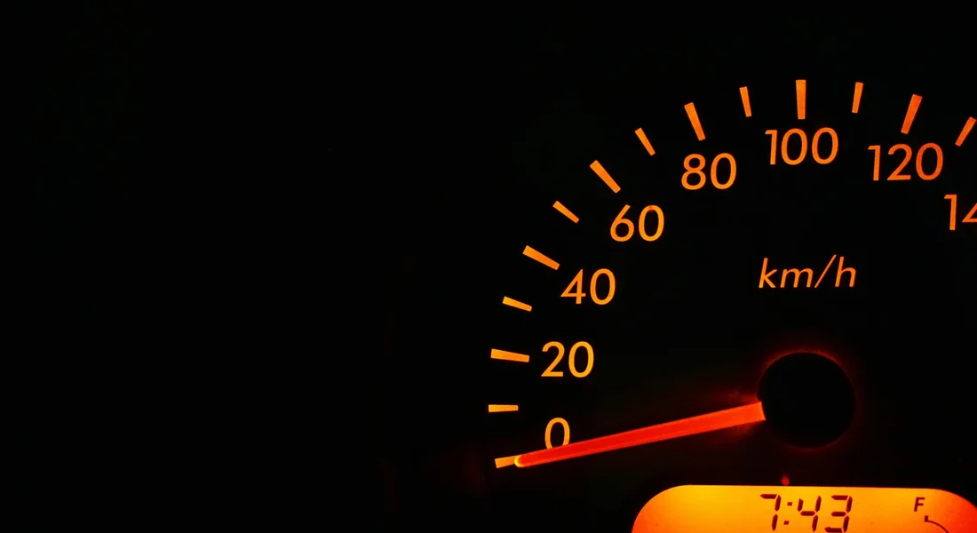 Close-up of a car dashboard speedometer illuminated in amber at night, with the needle near zero