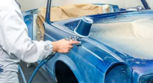 Technician wearing protective gear uses a spray gun on a blue car inside a paint booth.