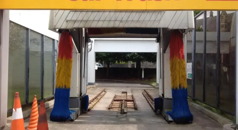 Entrance to an automatic car wash bay with a yellow overhead beam, red, yellow, and blue fabric-wrapped support columns, orange traffic cones, and a track for vehicles.