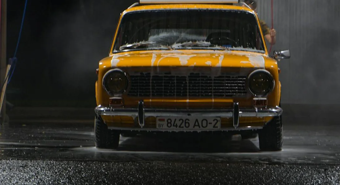A vintage yellow car being washed, with water spraying and droplets on the ground.