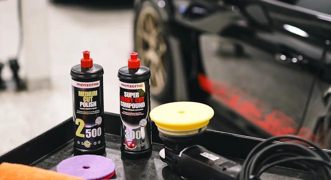 Car detailing setup on a workbench with wax polish bottles, applicator pads, and a polisher, with a car in the background