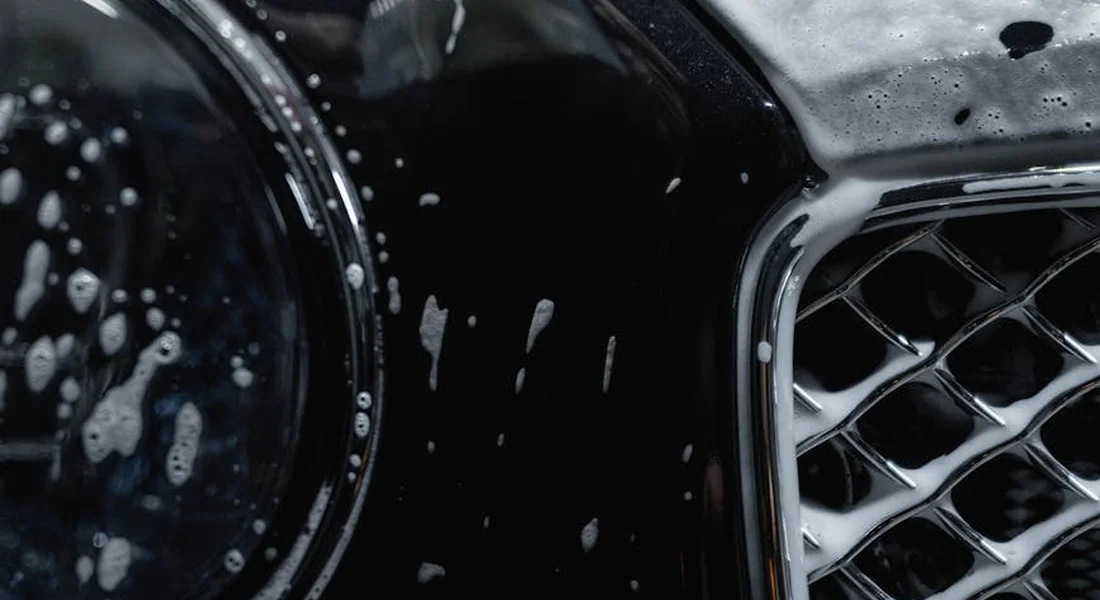 Close-up of a dark car’s front body with water droplets and soap splashes on the window area, highlighting common windshield cleaning challenges.