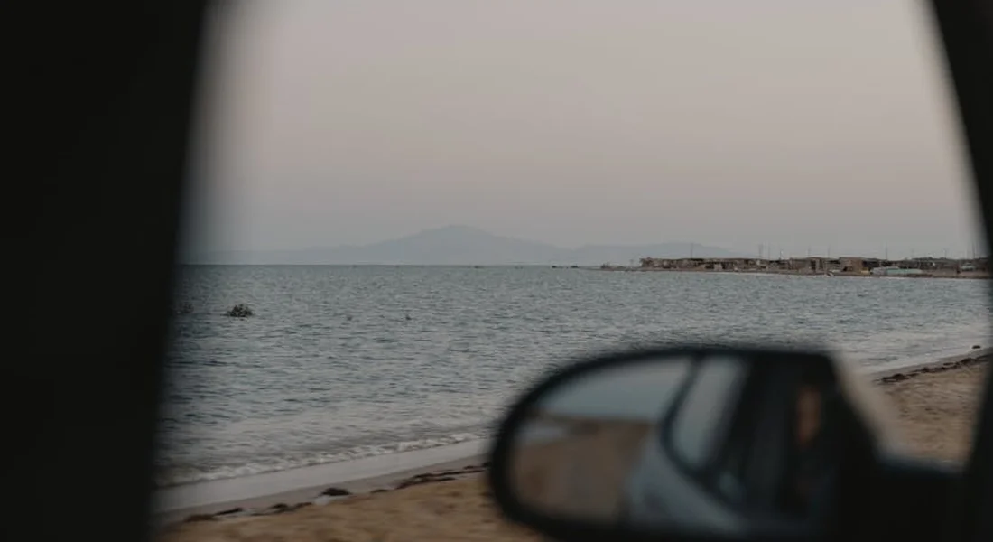 View through a car window at a calm seascape; the side mirror is in the foreground with water, beach, and distant mountains.