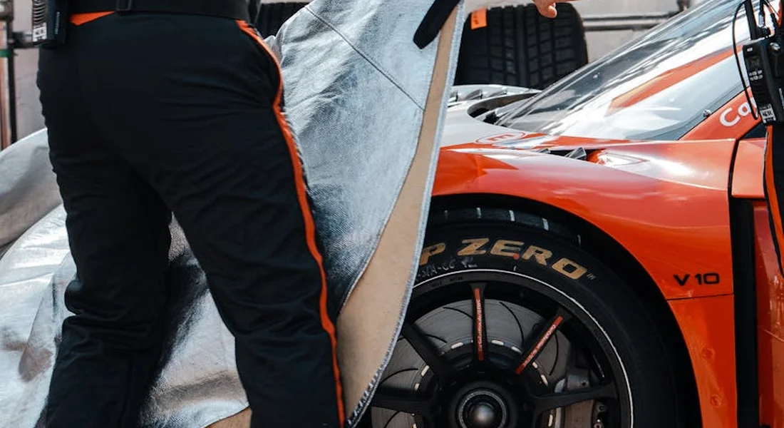 A technician applies a vinyl wrap to an orange race car, with the wrap sheet being laid over a body panel near the front wheel.