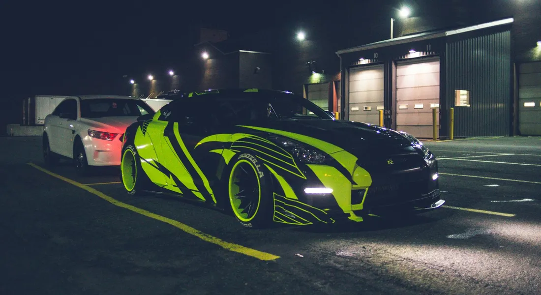 Nighttime scene of a neon-green wrapped sports car parked in a lot near a workshop, with another white car in the background.