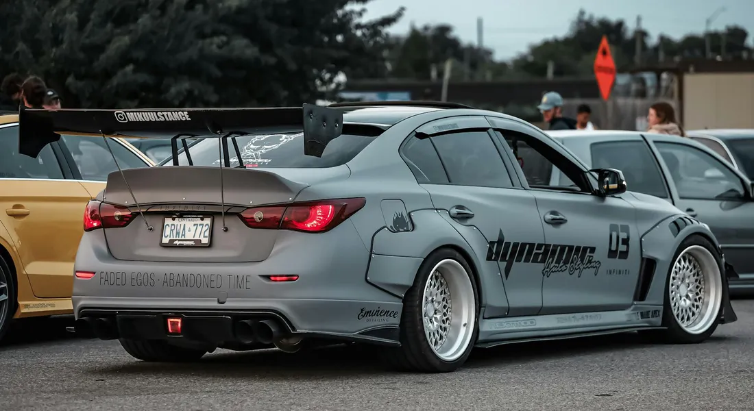 A grey modified sports coupe with a wide rear wing parked among other cars at a car meet, showcasing a complex body shape.