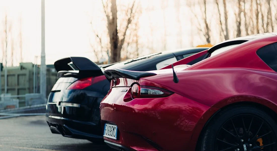 Two sports cars parked side by side in a lot, highlighting glossy red and black paint with rear windows and trim visible.