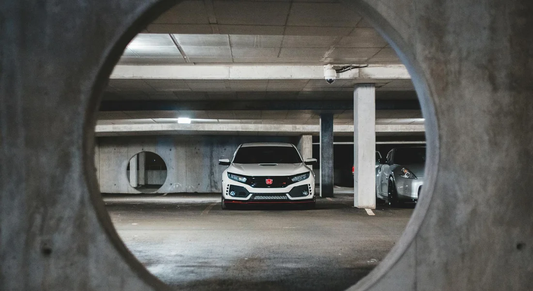 White car parked in a dim concrete parking garage, viewed through a circular opening in a concrete wall.