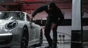 A technician in dark clothing detailing a white sports car in a workshop, with foam on the vehicle and equipment in the background.