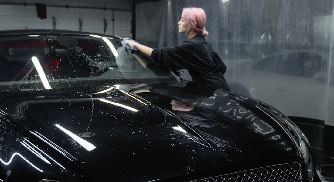 Person with pink hair wearing dark clothing washes a glossy black car in a workshop, wiping the windshield as water droplets scatter on the surface.