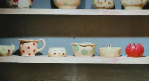 Row of glazed ceramic pots and bowls on wooden shelves against a blue wall.