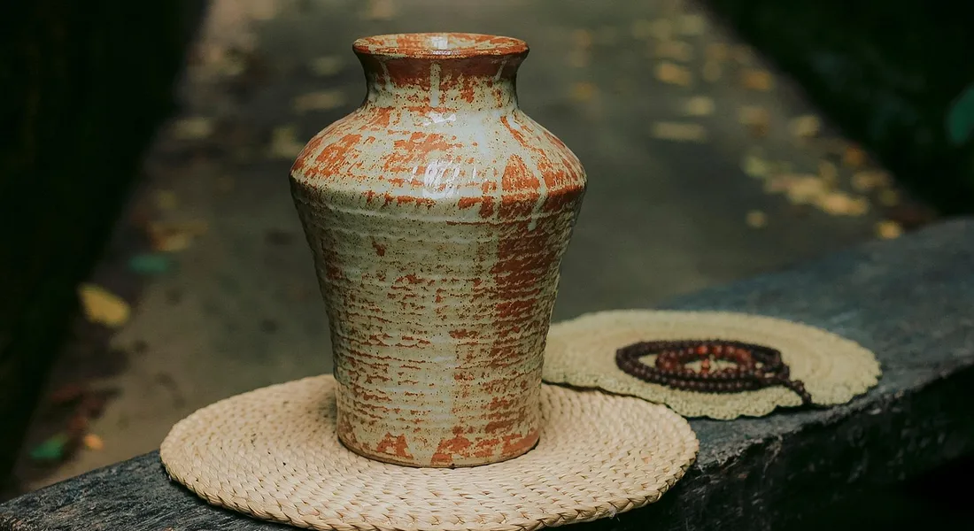 A rustic ceramic vase with a weathered glaze sits on a circular woven mat outdoors along a path.