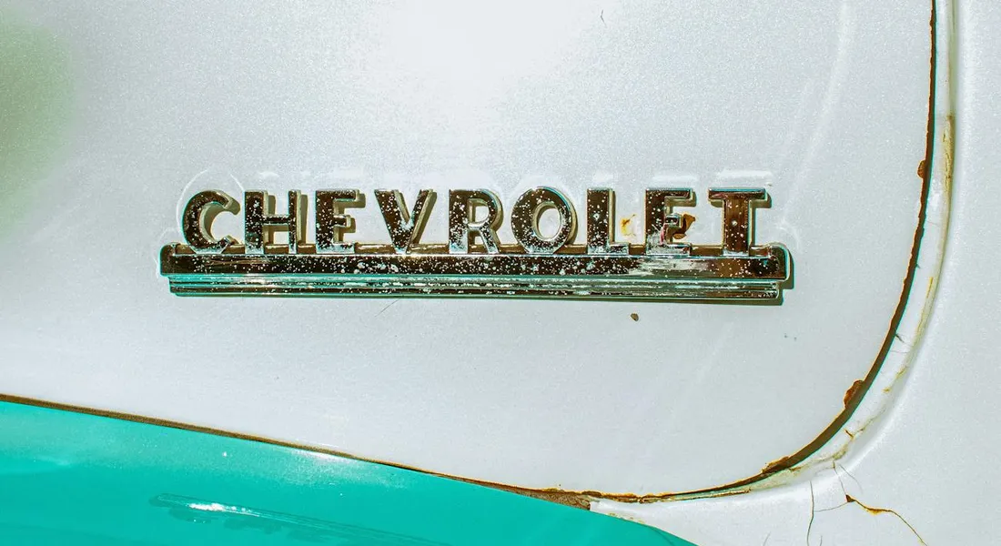Close-up of a vintage Chevrolet trunk with a chrome emblem, showing rust along the seam between white and turquoise paint.