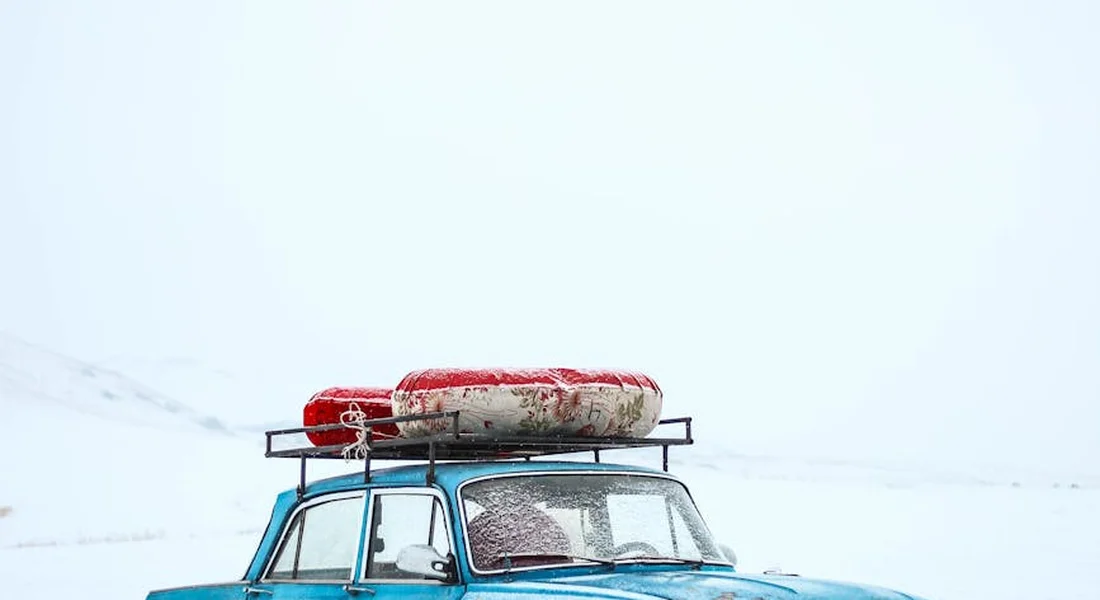 Blue vintage car with a red cargo bag on the roof rack in a snowy landscape.