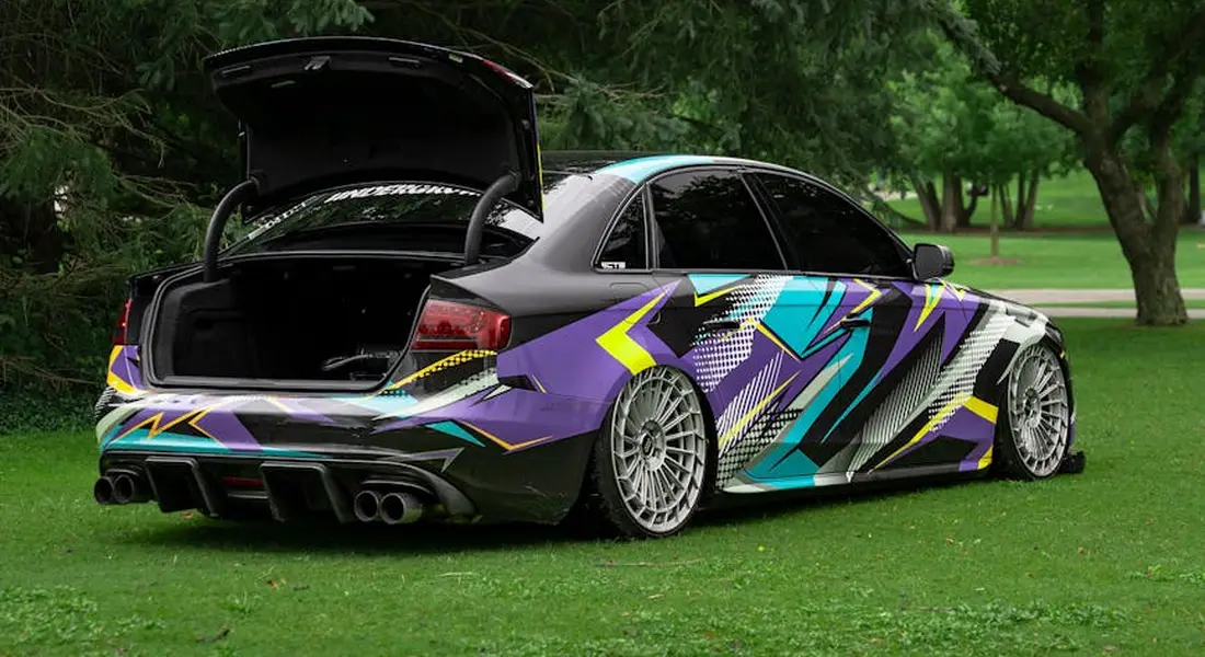A low-profile sports sedan with a bold, chrome-style geometric wrap parked on a grassy area, with the trunk open.