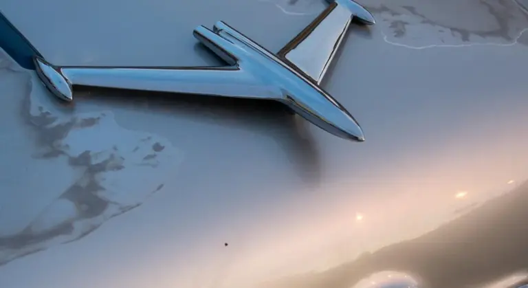 Close-up of a vintage car hood with a chrome emblem reflecting on a metallic silver surface.