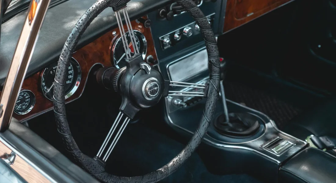 Classic car interior with a leather-wrapped steering wheel, chrome spokes, and a wood-grain dashboard.