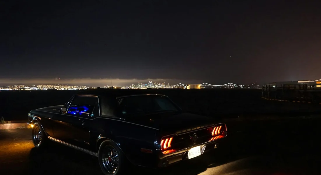 A classic black muscle car parked at night with city lights in the distance and a blue glow inside the interior.