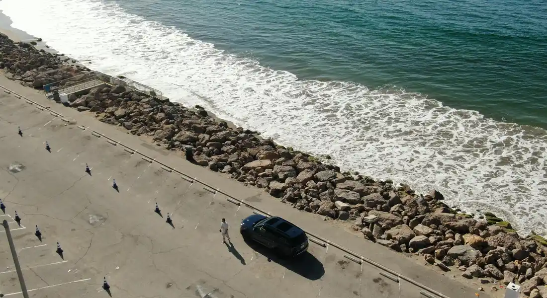 Aerial view of a car parked along a rocky shoreline with waves crashing onto the beach and a road running parallel to the coast.