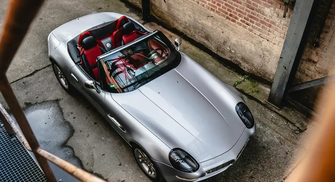 Top-down view of a silver convertible with the roof down and red interior, parked in an urban alley beside a brick wall.