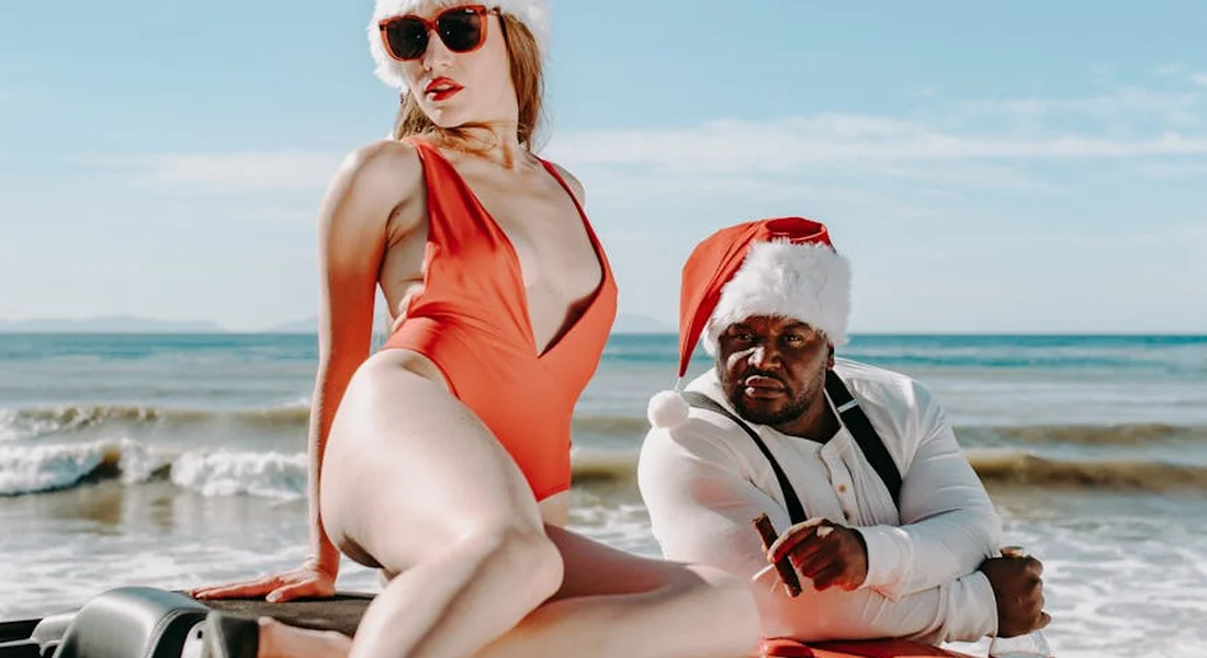 A woman in an orange swimsuit sits on the hood of a car on a sunny beach, wearing sunglasses, with a man in a Santa hat nearby.