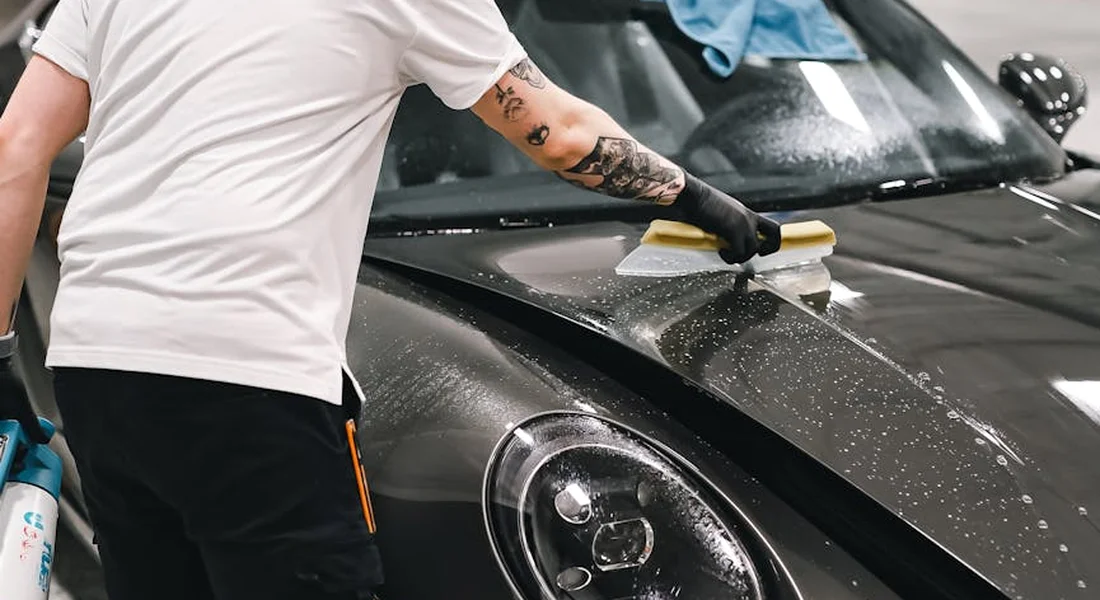 A detailer wearing a white shirt and gloves polishes a glossy black car hood with a handheld buffer.