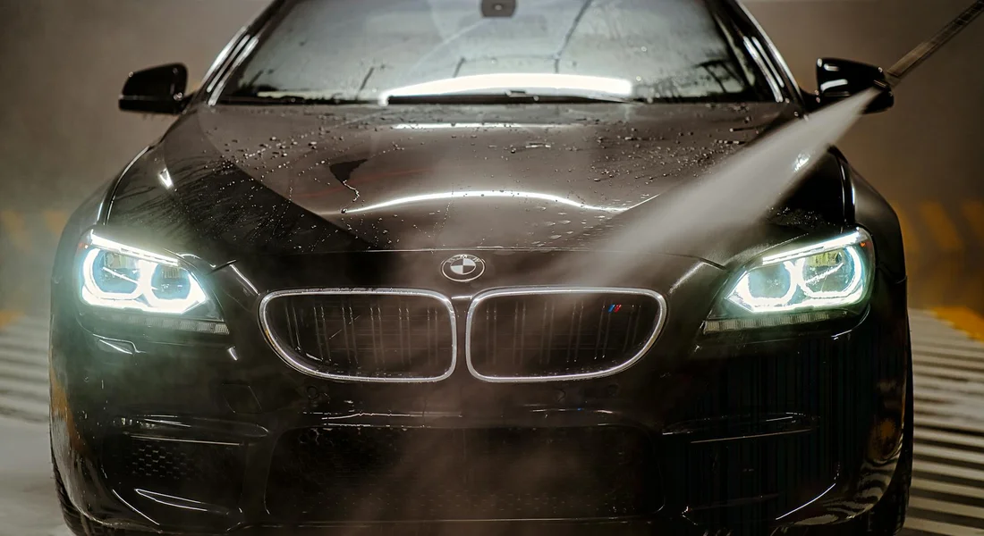 Front view of a black BMW being detailed, with a spray visible on the hood and illuminated headlights.