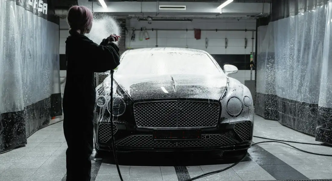 Detailer washing a black luxury car in a clean, indoor wash bay with a spray nozzle.