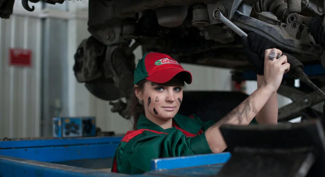 Automotive technician wearing a green uniform and red cap works under a raised car on a hydraulic lift, holding a tool.