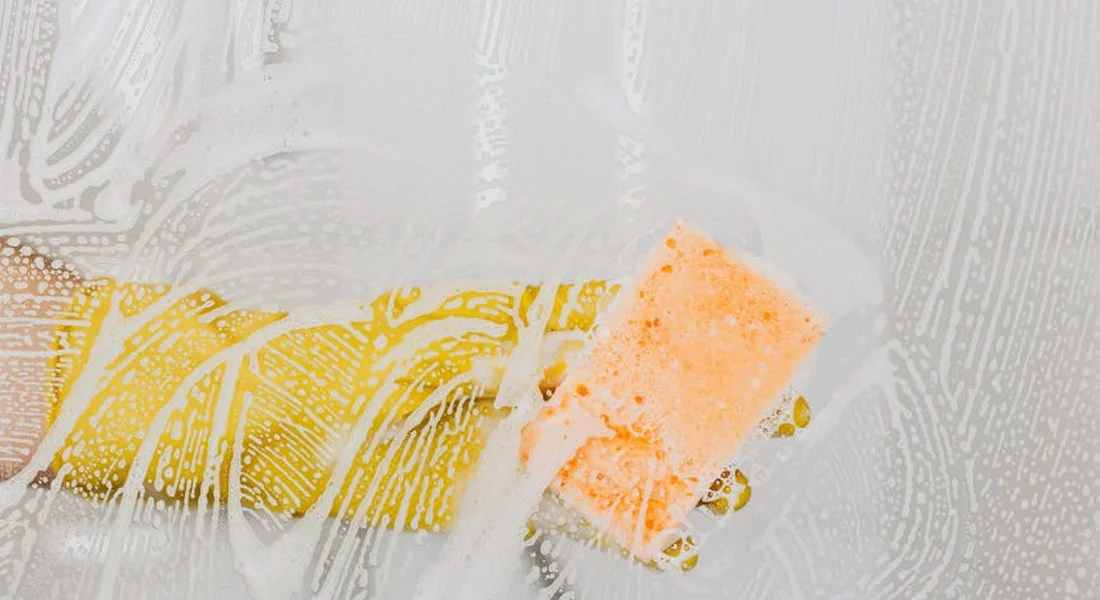 Close-up of a yellow sponge and scrub brush with soapy foam applying a cleaning in an engine bay.