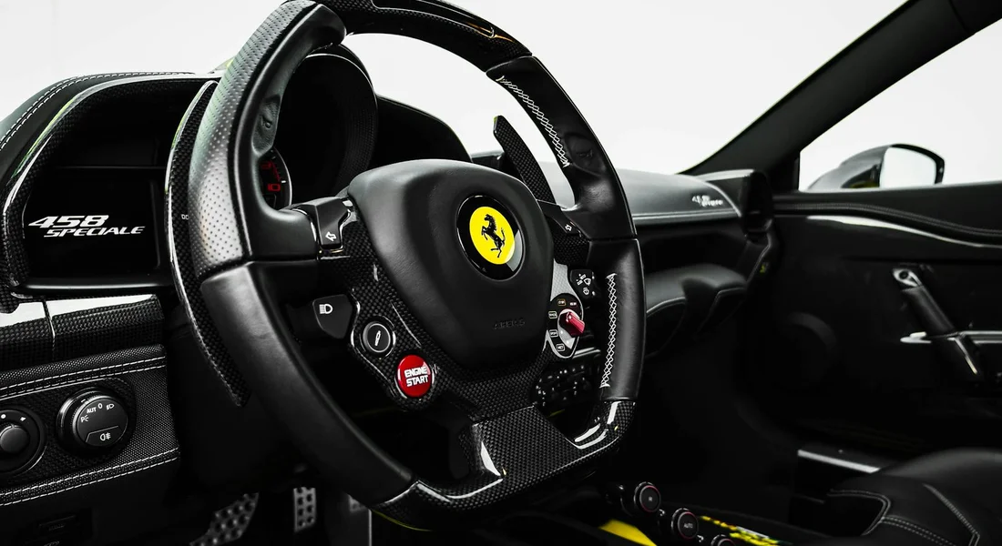 Close-up of a luxury car interior featuring a Ferrari steering wheel, carbon fiber dash, and red start/engine buttons.