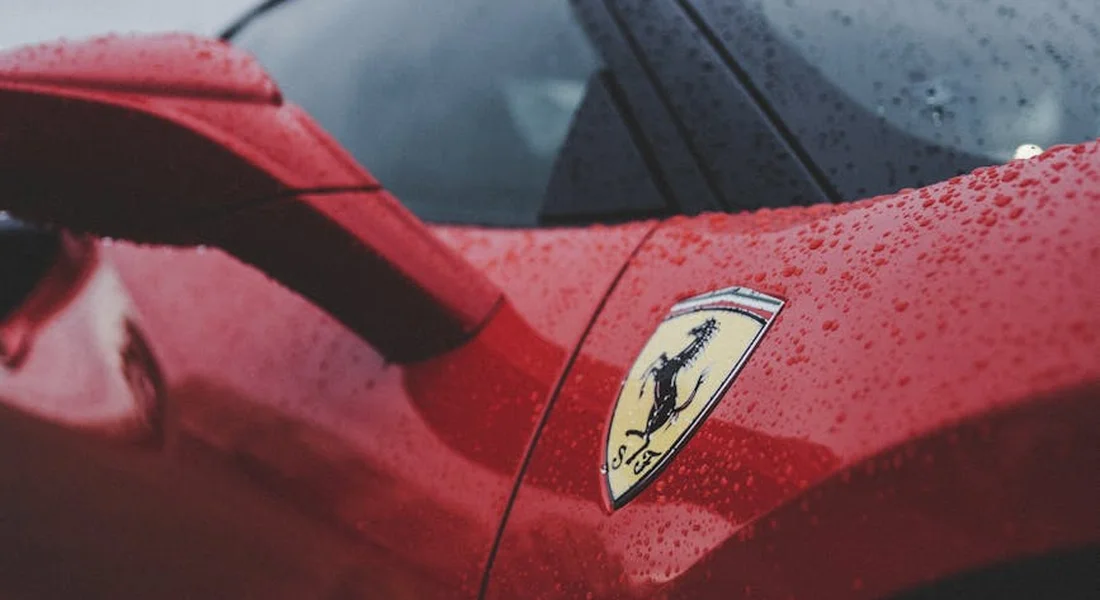 Close-up of a red Ferrari car showing the paint finish and a visible badge, with water droplets on the surface.