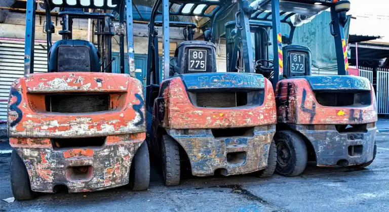 Three forklifts with chipped orange and blue paint parked side by side in a dusty lot, showing weathered finishes and scratches.