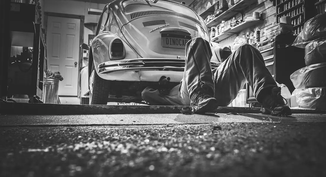 A DIY enthusiast works under a classic car in a cluttered home garage, preparing to touch up paint and blend edges.