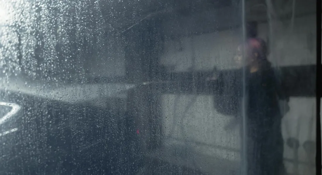 Close-up of a wet car window with water droplets and a blurred reflection of a person taking a photo