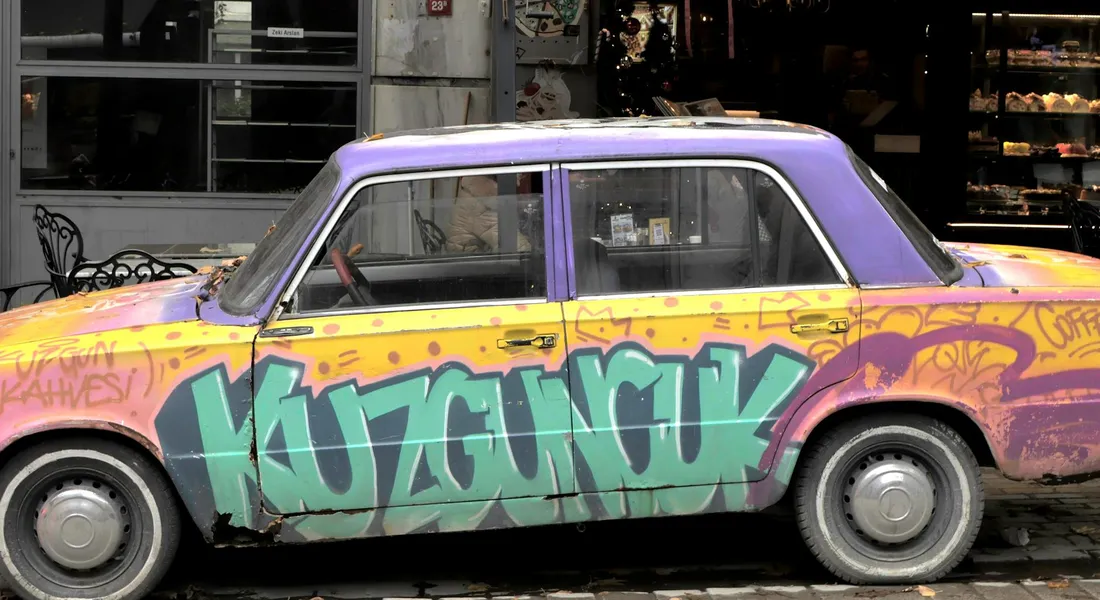 A vintage car with purple and yellow graffiti along its side, parked beside a storefront.