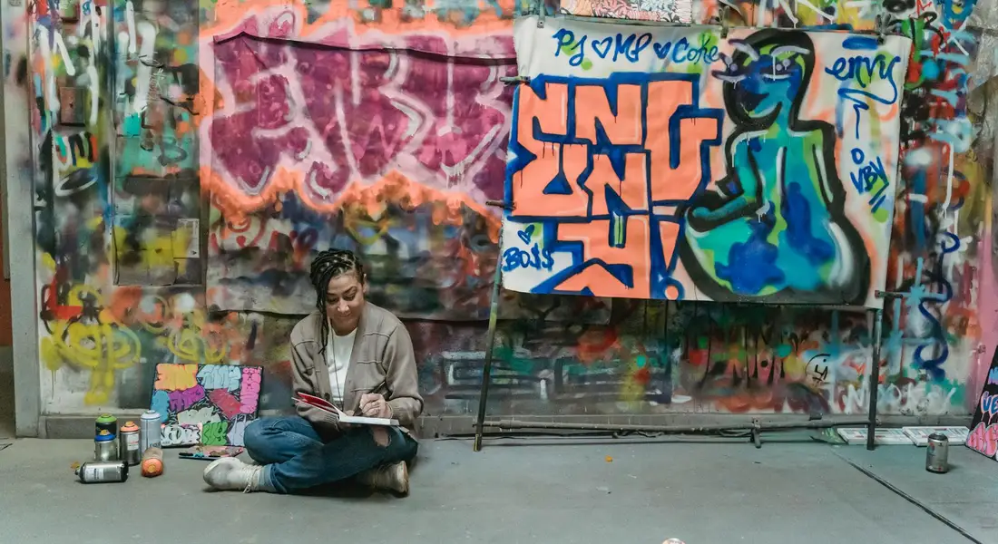 A graffiti artist sits cross-legged on the ground, sketching while surrounded by spray cans in front of a colorful mural on a wall.