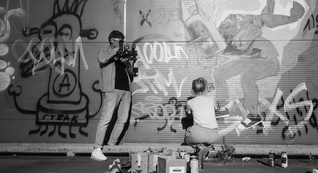 A photographer films a graffiti-covered wall as spray paint cans sit on the ground.