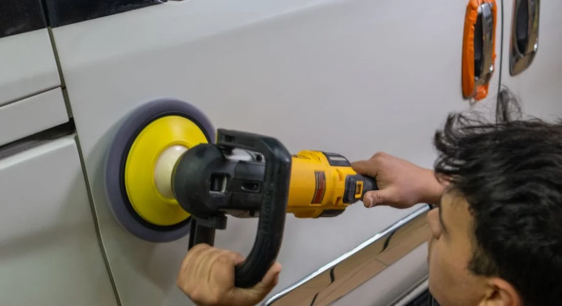 A person operating a yellow polishing tool to remove gum from a car's paint near the exterior trim.