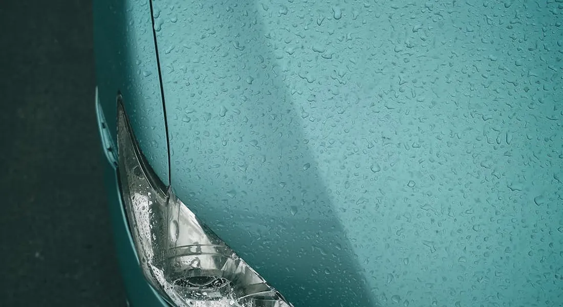 Close-up of a teal car hood covered in water droplets, with a portion of the headlight visible
